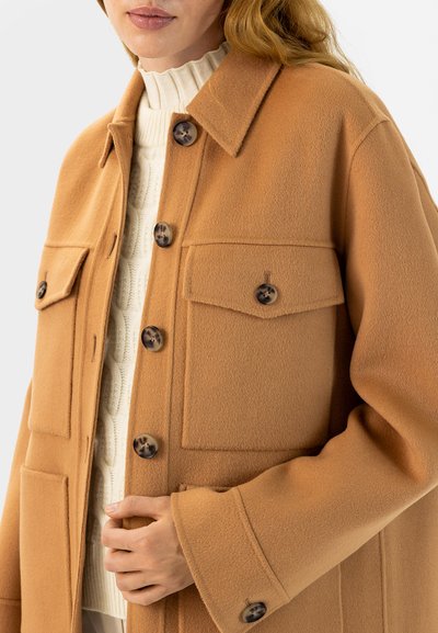 Kamelhaarfarbener Wollmantel mit Kragen, Taschen vorne und großen braunen Knöpfen. Strukturierter Stoff und lässige Passform. Auffällige Kombination mit einem gestrickten Oberteil.