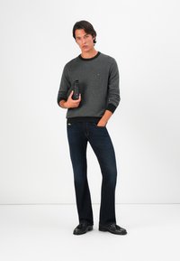 Grauer Sweatshirt mit schwarzem Rippstrickbund, getragen mit dunkelblauen Jeans und schwarzen Slippers, gehalten mit einer schwarzen Accessoires-Tasche.
