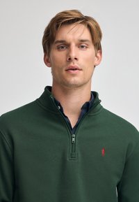 Pullover verde realizzato in tessuto morbido, con collo a zip a quarto, logo rosso ricamato sul petto e colletto strutturato.