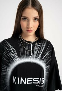 Młoda kobieta z długimi brązowymi włosami, ubrana w czarną koszulę z białym wzorem słonecznym i tekstem "KINESIS subconscious", ozdobiona srebrnymi naszyjnikami.
