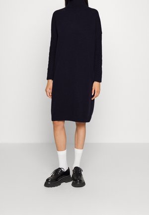 Osoba ubrana w granatową dzianinową sukienkę z długim rękawem, białe skarpetki za kostkę i czarne masywne buty na sznurowadła, stojąca na jednolitej podłodze.