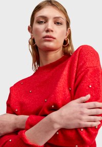 Pull en tricot rouge avec un col rond, orné de clous métalliques et de poignets côtelés. Texture lisse, porté avec des boucles d'oreilles surdimensionnées.