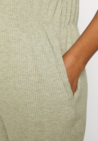 Macacão em verde claro com textura ribada, apresentando bolsos laterais e uma faixa elástica na cintura para um ajuste confortável. O tecido tem uma aparência macia e texturizada.