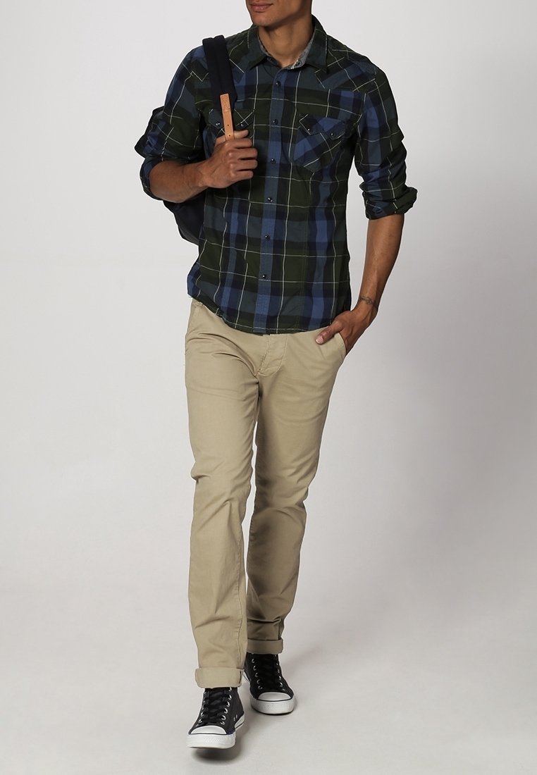 Homme marchant avec la main dans la poche, portant une chemise à carreaux verte et bleue, un pantalon beige, des baskets noires et portant un sac à dos noir sur une épaule.