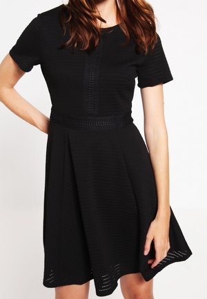 Svart klänning med kort ärm, texturerad yta, figursydd midja och utsvängd kjol; har en subtil vävd detalj vid midjan och fållen.