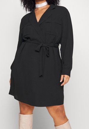 Kvinde iført en sort, langærmet bæltekjole med brystlommer og en sølvfarvet choker, kombineret med beige, knælange støvler.