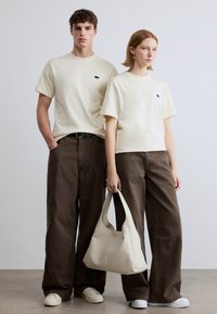 Μπεζ μπλουζάκια με μαύρο λογότυπο, χαλαρά καφέ παντελόνια και λευκή τσάντα χόμπο. Τα μοντέλα ποζάρουν με ουδέτερο φόντο και casual λευκά αθλητικά παπούτσια.