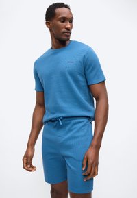 Άνδρας που φοράει μπλε κοντομάνικο πουκάμισο με υφή και κοντό παντελόνι που ταιριάζει, κοιτάζοντας προς τα κάτω αριστερά, στέκεται μπροστά σε μια απλή φωτεινή επιφάνεια.