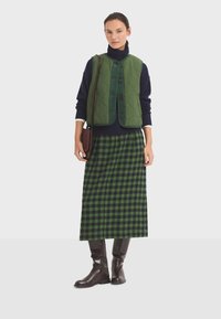 Grüne Steppweste über einem marineblauen Rollkragenpullover, kombiniert mit einem langen karierter Rock in Grün und Marineblau und braunen knielangen Stiefeln. Braune Umhängetasche.