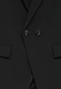 Gros plan d'un blazer noir à double boutonnage avec deux boutons noirs brillants et des poches à rabat de chaque côté.