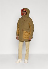 Ocieplana kurtka parka w kolorze oliwkowym i beżowym, z wysokim kołnierzem, dużym kapturem i dużą przednią kieszenią z pikowanymi akcentami. Białe trampki.