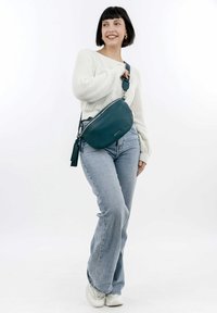 Zelená kožená crossbody taška s zaoblen�ým tvarem, texturovaným povrchem, stříbrným zipem a třásněmi. Model ji nosí s volnými džíny a svetrem.