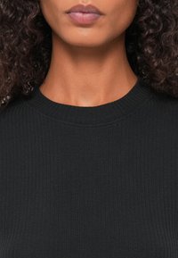 Svart ribbestrikket topp med høy hals. Teksturen er glatt med vertikale linjer, noe som fremhever en figursydd design.