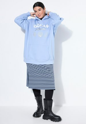 Nainen pukeutunut vaaleansiniseen oversize-huppariin, jossa lukee "OCEAN please" ja simpukkakuviot, raidallinen hame, mustat leggingsit ja paksut mustat saappaat.