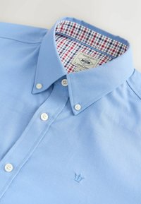 Šviesiai mėlynas marškinis su apykakle, su mygtukų užsegimu, baltais mygtukais ir kontrastiniu patikros raštu apykaklės viduje. Išsiuvinėtas simbolis ant krūtinės.