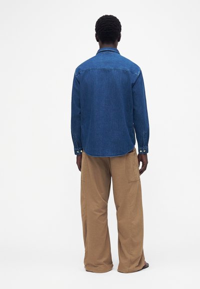 Homme portant une chemise en jean bleue et un pantalon beige à jambes larges, debout, dos tourné, sur un fond clair uni.