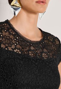 Schwarzes Spitzentop mit floraler Stickerei, Rundhalsausschnitt mit gewelltem Besatz und einem taillierten unteren Abschnitt. Große geometrische Ohrringe aus Silber und Gold.