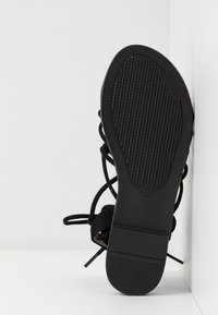 Semelle d'une sandale noire à brides avec une semelle texturée, taille 37, appuyée contre un mur d'angle blanc sur une surface lisse.