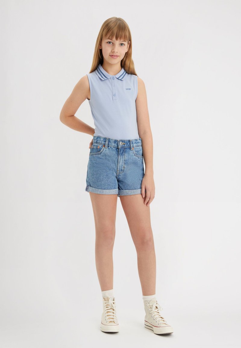 Polo sin mangas de color azul claro con un cuello bicolor, combinado con shorts de mezclilla de tiro alto. Zapatillas blancas de caña alta completan el atuendo.