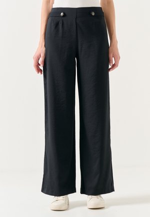 Pantaloni neri a gamba larga con una texture liscia, dotati di due tasche anteriori e dettagli con bottoni in argento in vita. Abbinali a delle sneakers bianche.