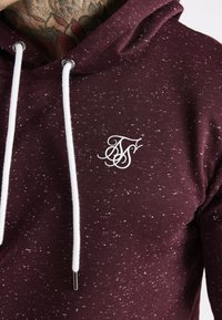 Primo piano di una felpa con cappuccio color borgogna con macchie bianche, coulisse bianche e un logo stilizzato bianco sul petto, che mostra parzialmente un collo tatuato.