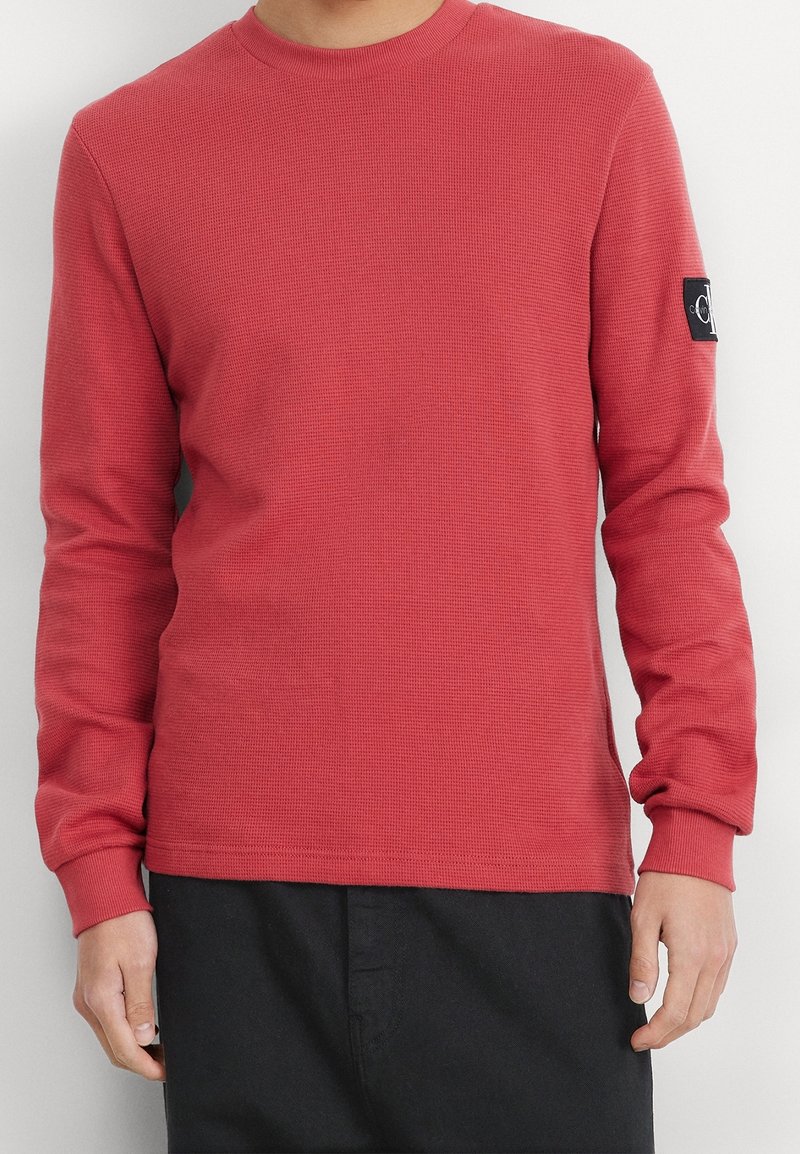 Röd, långärmad, texturerad crew neck sweatshirt med en svart logotyp patch på vänster ärm och ribbad mudd. Svarta byxor synliga nedanför.