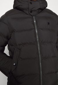 Černá péřová bunda s vysokým límcem, hladkým povrchem, předním zipem a cvoky. Má boční kapsy a nenápadný detail loga.