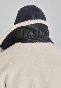 Jachetă albă din blană de oaie cu guler din piele neagră, având branding embosat. Gluga este de culoare bleu marin, evidențiind contrastul dintre cele două materiale.
