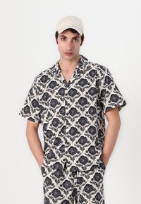 Κοντομάνικο πουκάμισο από κρεμ ύφασμα με ναυτικό λουλουδάτο σχέδιο, σε χαλαρή γραμμή και σχέδιο με γιακαδάκι κάμπινγκ. Διαθέσιμα και παντελόνια σε αντίστοιχο σχέδιο.