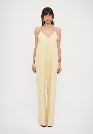 Donna in piedi di fronte che indossa una tuta senza maniche, di colore giallo pallido, con spalline sottili e gambe larghe, con tatuaggi visibili sulle braccia e orecchini.