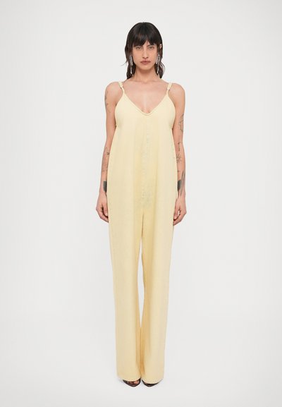 Femme debout, de face, portant une combinaison ample sans manches jaune pâle avec fines bretelles et jambes larges, avec des tatouages visibles sur les bras et des boucles d'oreilles.