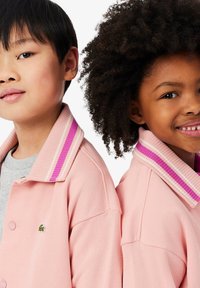 Růžová bunda s proužkovanými detaily límce v odstínech světle růžové a fialové. Disponuje zapínáním na knoflíky a malým logem na levé straně.