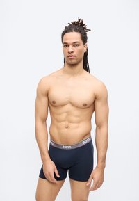 Μυώδης άνδρας με ντραντλόκς, που φοράει σκούρα μπλε εσώρουχα BOSS, στέκεται μπροστά σε ένα απλό ανοιχτό γκρι φόντο.