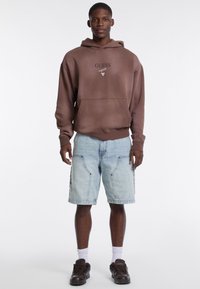 Felpa marrone con tasca frontale e logo del marchio, abbinata a pantaloni shorts in denim azzurro chiaro e sneakers marroni scuri. Calze bianche visibili.
