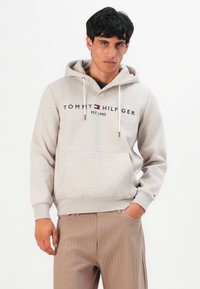 Jonge man in een beige Tommy Hilfiger hoodie met logo en koorden, gecombineerd met lichtbruine gestreepte broeken, staand tegen een witte achtergrond.