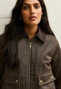 Vrouw met lang zwart haar, gekleed in een bruine zip-up jas met klepzakken en een geribde kraag over een beige trui.