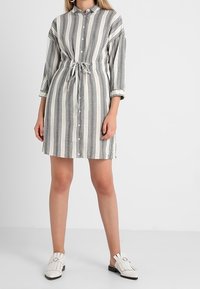 Modelka ubrana w pasiowaną sukienkę zapinaną na guziki z przewiązaniem w talii, zestawioną z białymi butami typu slip-on, na jednolitym tle.