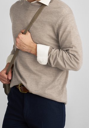 Homme portant un pull beige sur une chemise blanche, tenant la sangle olive d'un sac à bandoulière, vêtu d'un pantalon bleu marine avec une ceinture marron.