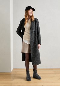 Svartvit prickig yllemantel, krämfärgad ribbstickad tröja, beige kjol, svarta strumpbyxor och svarta snörkängor, bärs med en svart keps.