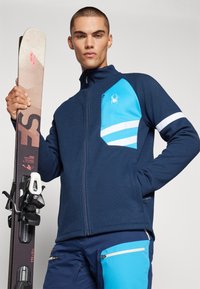 Veste zippée navy avec une finition texturée, présentant une poche poitrine bleu clair, des rayures blanches sur les manches, et un logo d'araignée. Équipement de ski en main.