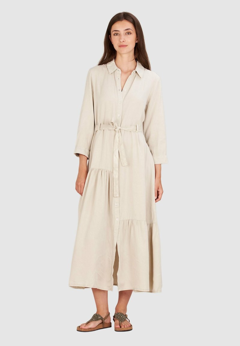 Długa beżowa sukienka koszulowa z guzikiem na przodzie, paskiem w talii i warstwowym dołem. Wykonana z lekkiego materiału o gładkiej fakturze.