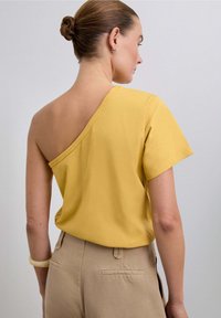 Haut asymétrique jaune en tissu doux, avec une manche courte d'un côté et un dos ouvert, associé à un pantalon beige.