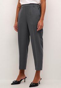 Pantalons gris ajustés avec taille haute, devant plissé et jambes raccourcies et fuselées. Le tissu a une texture lisse, associés à des mules noires à bout pointu.