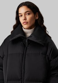 Chaqueta puffer negra con un cuello alto y estructurado; presenta una cremallera frontal y textura acolchada. Lleva puesta sobre un suéter de canalé color crema.
