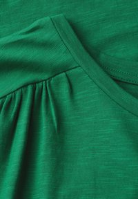 Green fabric close-up showing gathered sleeve detail and stitching on soft, lightweight knit material.