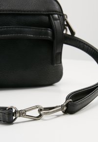 Mustapintainen nahkainen laukku irrotettavalla hihnalla, jossa on hopeinen metalliklipsi ja vetoketjullinen kolo edessä.