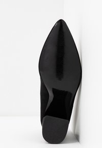 Semelle de chaussure noire à bout pointu avec talon texturé placée verticalement contre un mur d'angle blanc.