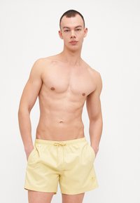 Κίτρινα μαγιό φτιαγμένα από ελαφρύ ύφασμα, διαθέτοντας ελαστική μέση με κορδόνι και μια μικρή λεπτομέρεια λογότυπου στην αριστερή πλευρά.