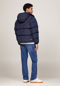 Giubbotto puffa blu con cappuccio, caratterizzato da un design a trapunta, polsini elasticizzati e dettaglio del logo sull'orlo, abbinato a jeans blu e sneakers bianche.
