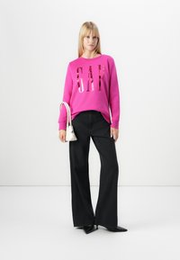 Fuchsia genser med stort reflekterende "GAP"-logo, kombinert med vide svarte jeans. Modellen holder en liten hvit håndveske.